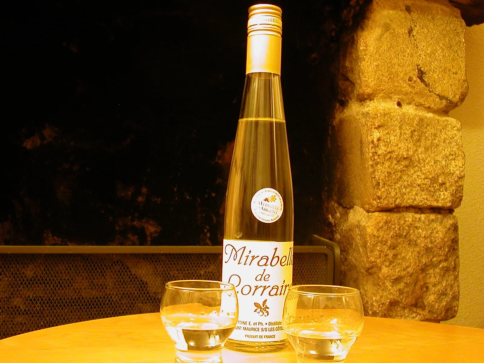Une bouteille d’eau-de-vie de mirabelle de Lorraine et deux petits verres sur une table, devant un mur de pierre