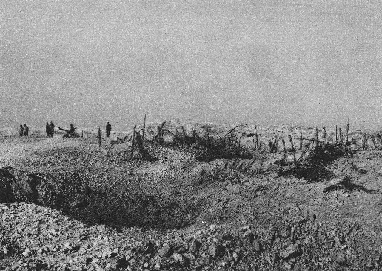 Photographie ancienne en noir et blanc : un vaste trou d’obus au premier plan, des piquets et des barbelés arrachés, quelques silhouettes au loin sur un sol nu