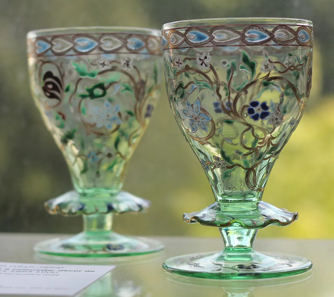 Deux verres à pied d’Émile Gallé en verre vert clair, décorés de fleurs émaillées bleues et de rinceaux dorés